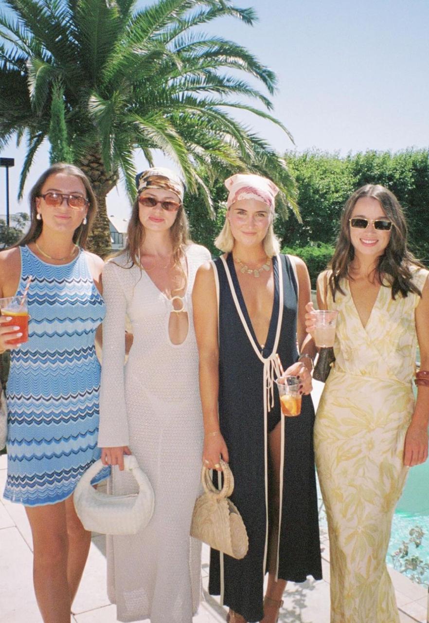 Women at a sunny pool wearing elegant summer dresses, sunglasses, headscarves, and resort accessories